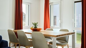 A dining area with a wooden table and four chairs, large windows with red curtains, and a bowl of fruit on the table. - Beach House in France near the Coast (Berck-Sur-Mer)