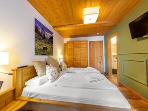 A hotel room with a large bed, wooden headboard, a mounted TV, and a painting of a mountain landscape. - Penthouse Apartment with Mountain Views (Neukirchen am Großvenediger)