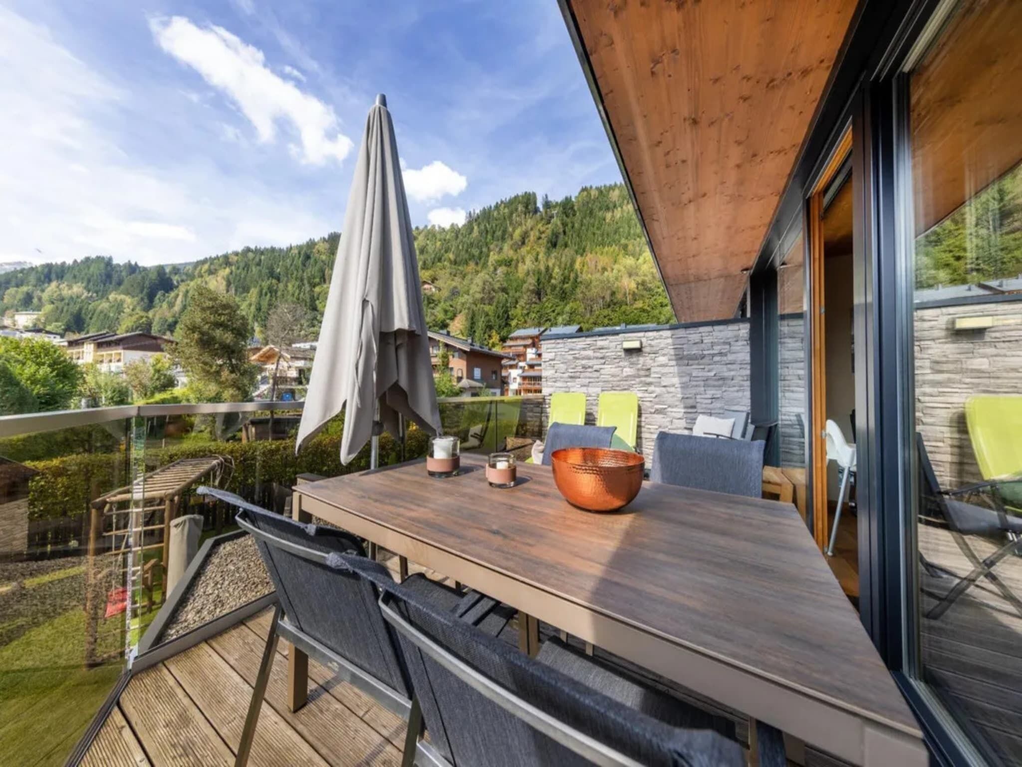 Terraza de madera con una mesa, sillas y barandilla de cristal con vistas a un paisaje montañoso.