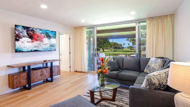 A modern living room with a large flat-screen TV, a glass coffee table, and a grey sofa with patterned cushions.