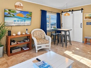 A cozy living area with a television displaying a tropical beach scene, a wicker chair, a wooden side table with decorative items, and a dining area with a table and bar stools. - Direct Oceanfront and Boardwalk Condo on 12th St. with Indoor Pool! Decatur House 505 (Ocean City)