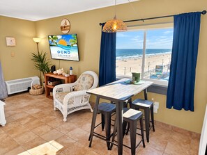 A room with a view of the beach, a table with stools, a wicker chair, a TV on the wall, and a hanging light fixture. - Direct Oceanfront and Boardwalk Condo on 12th St. with Indoor Pool! Decatur House 505 (Ocean City)