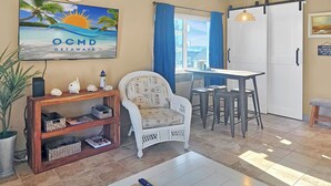 A living room with a beach-themed painting, a white wicker chair, a small table, and a coffee table.