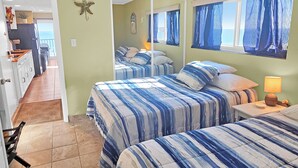 A bedroom with two beds, a nightstand, a lamp, a mirror, and a view of the kitchen and balcony.