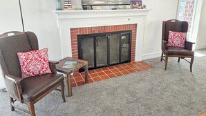 A living room with a fireplace, two armchairs with red and white patterned cushions, a small table with a book, and a television displaying 'OCMD GETAWAYS'.