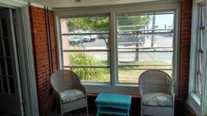 Une pièce d’angle avec deux fauteuils en osier et une petite table, ainsi qu’une grande fenêtre donnant sur la rue et les voitures garées.