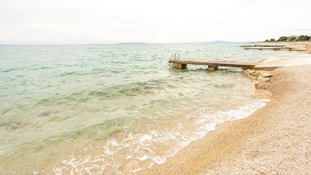 Una playa con un muelle de hormigón que se adentra en aguas tranquilas, costa de guijarros y colinas a lo lejos.