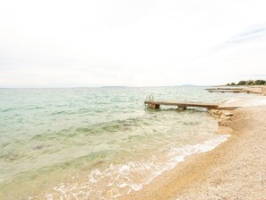 A beach with a concrete pier extending into calm waters, pebbly shore, and distant hills.