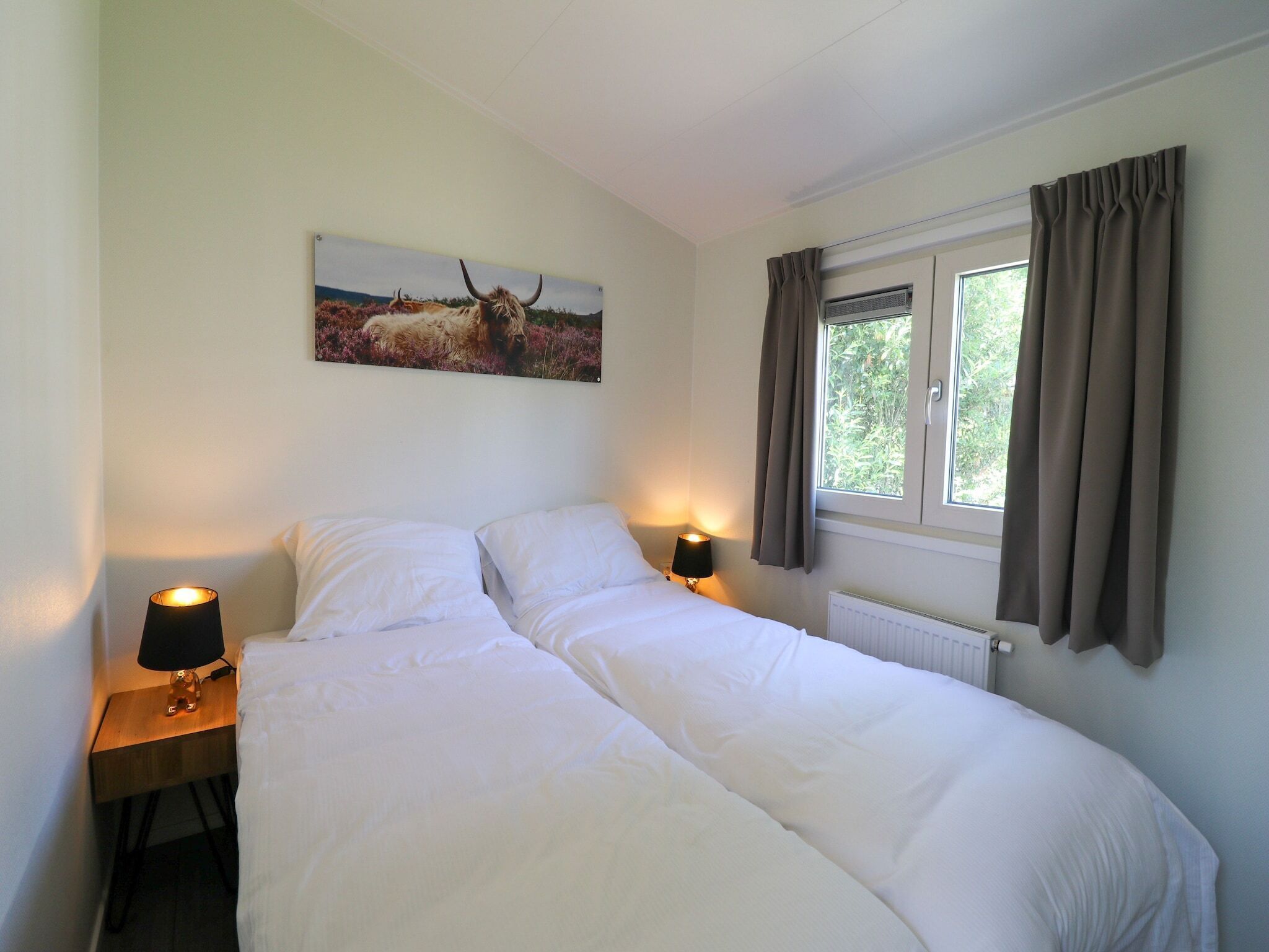 A bedroom with a bed, two bedside lamps, a window with curtains, and a painting on the wall.