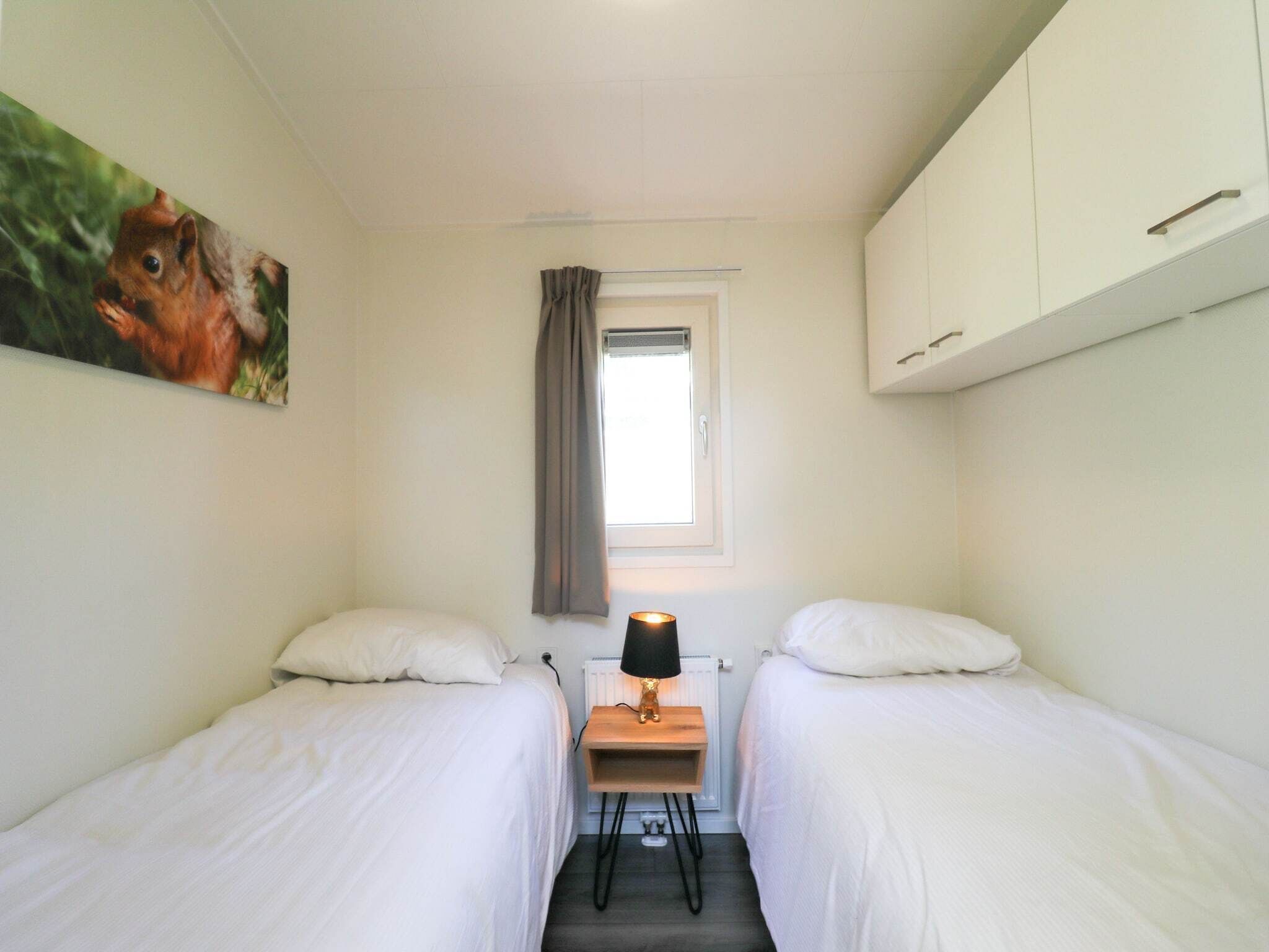 A small, clean room with two single beds, a nightstand with a lamp, a window with curtains, and a painting on the wall.