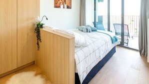 Een slaapkamer met een houten garderobe, een bed met witte beddengoed, een kleine plant en een balkon met uitzicht.