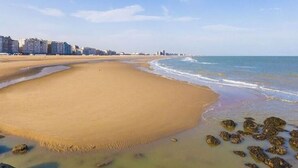 Een breed zandstrand met een kalme zee, een heldere lucht en gebouwen op de achtergrond.