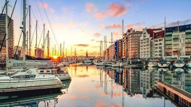 Een jachthaven met verschillende boten afgemeerd, omringd door gebouwen, met een zonsondergang op de achtergrond.