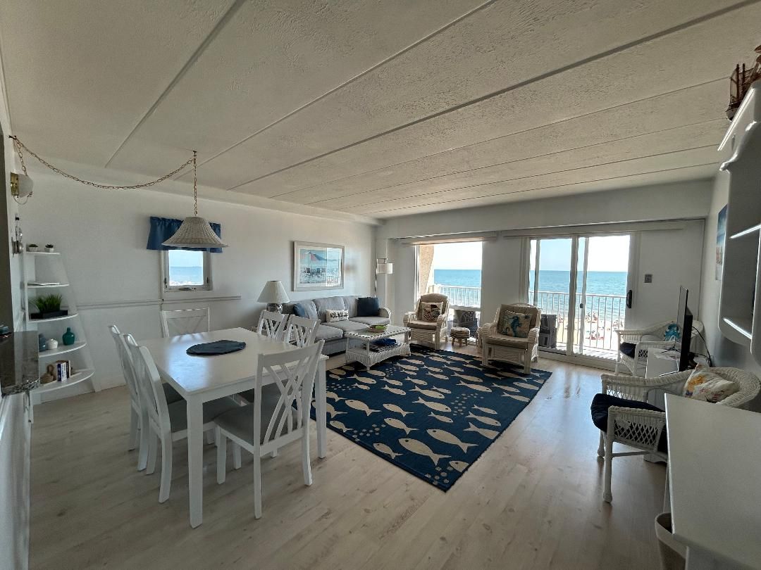 Un vaste espace de vie comprenant une table à manger, des meubles blancs, un tapis à motifs bleus et une vue sur la mer grâce à de grandes fenêtres.