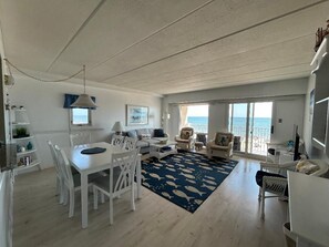 A spacious living area with a dining table, white furniture, a blue patterned rug, and a view of the sea through large windows. - OCEAN FRONT CONDO.  ENJOY YOUR MORNING COFFEE SITTING ON YOUR PRIVATE BALCONY WATCHING THE SUNRISE. (Rehoboth Beach)