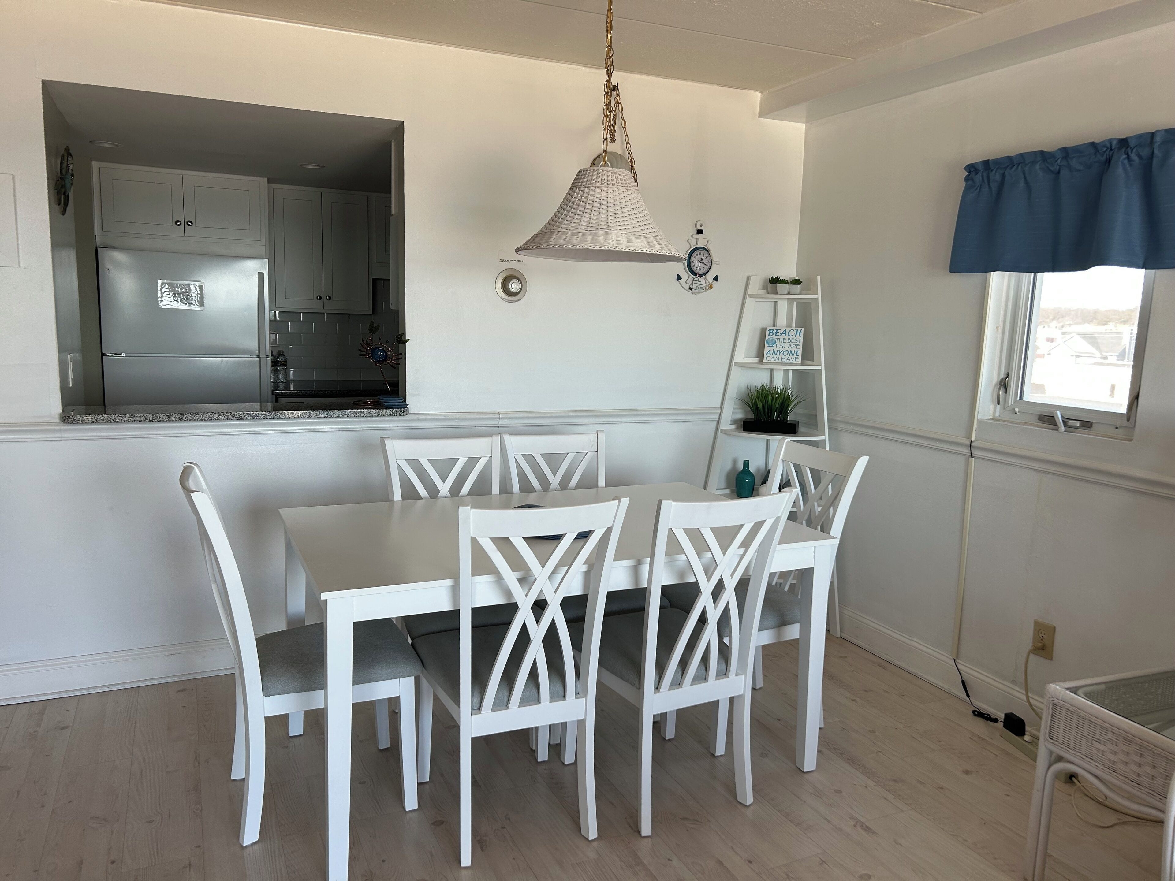 Een eethoek met een witte tafel en stoelen, een hanglamp, een keuken die door een open deur zichtbaar is, en een raam met een blauwe gordijnboog.