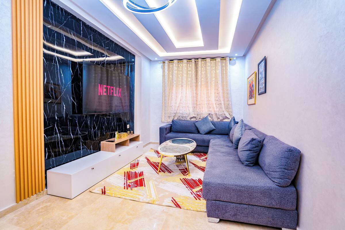 Uma sala de estar moderna com um sofá cinza, um tapete estampado e uma janela grande com a inscrição Netflix.