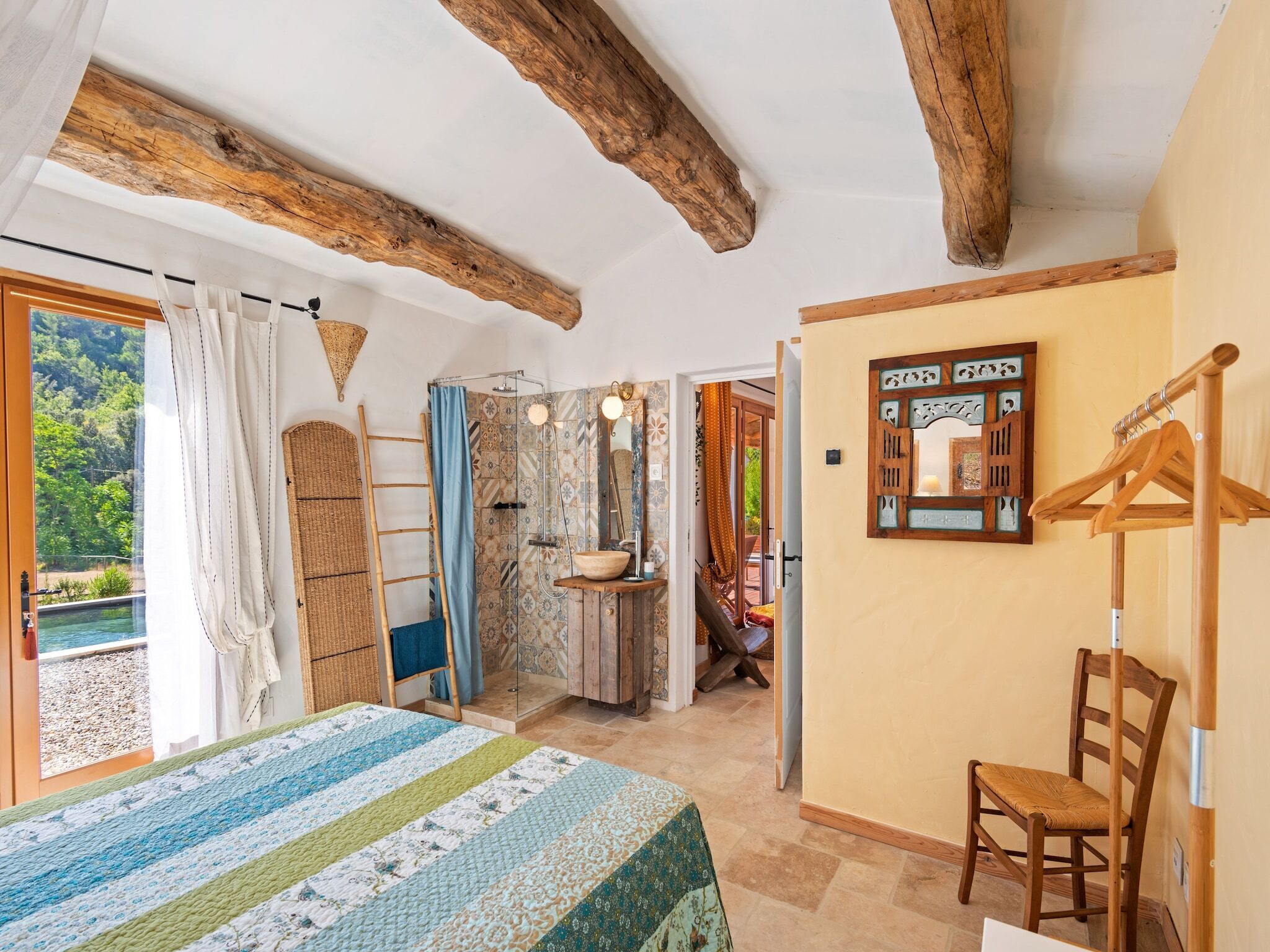 A bedroom with a bed, a wooden chair, a bathroom with a shower, and a view of a forest.