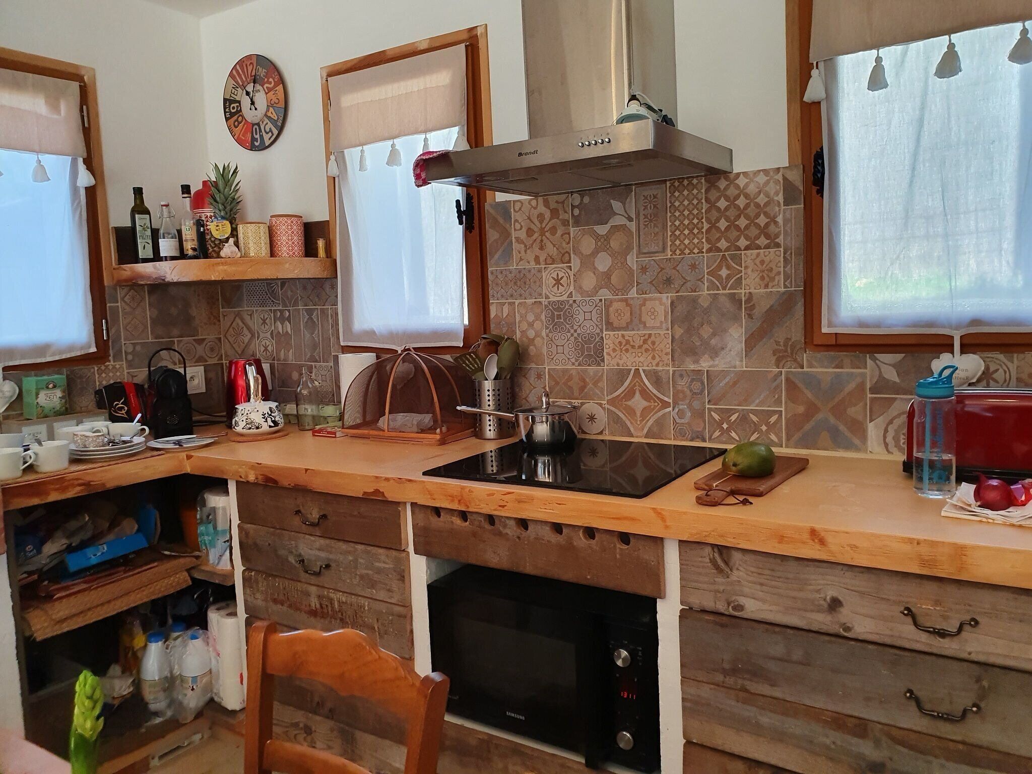 A kitchen with a stove, oven, and a window with curtains. There are various kitchen items on the counter, including a kettle, a teapot, and a water bottle.