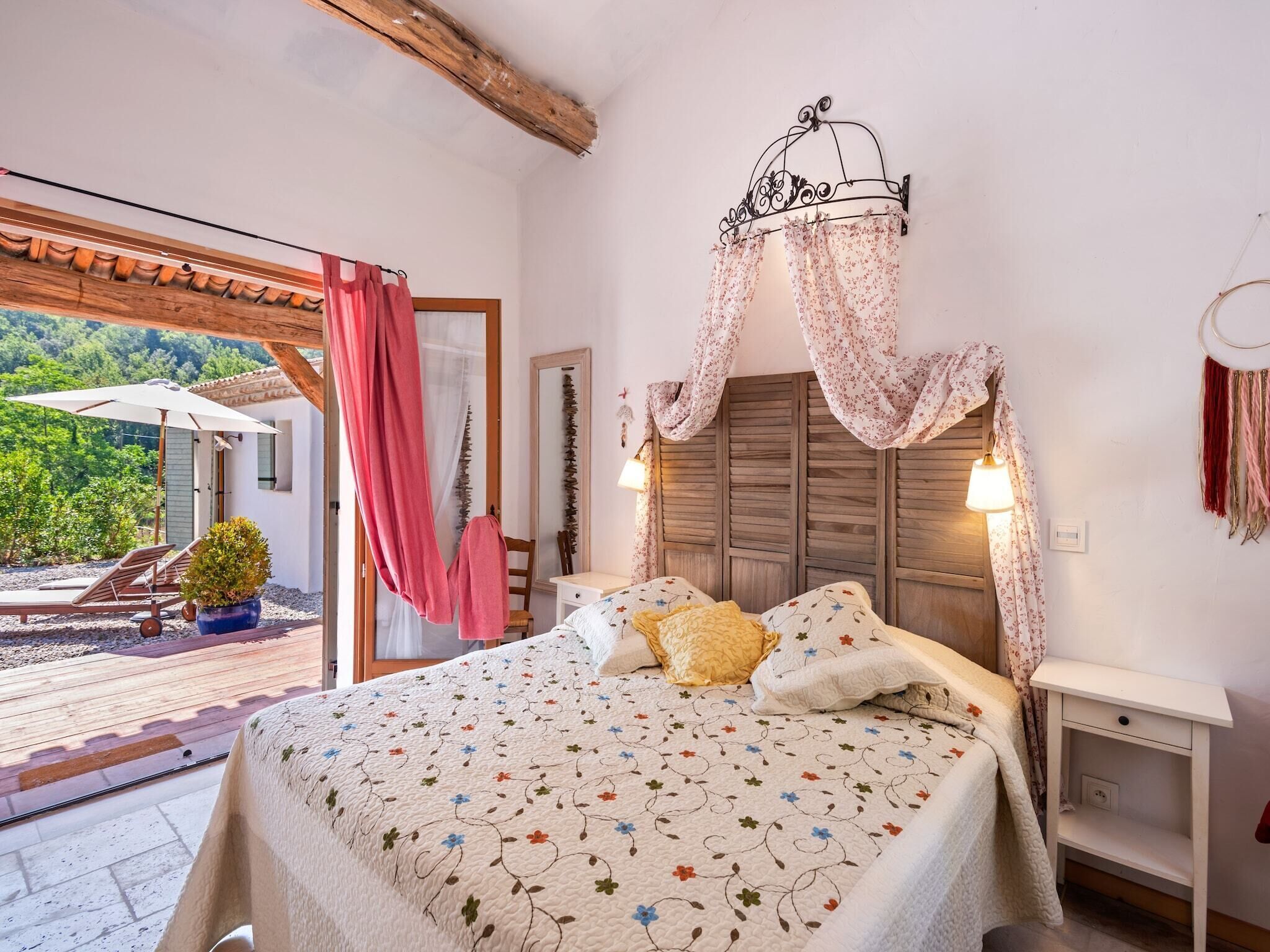 A bedroom with a bed, a wooden headboard, a nightstand, a window with a view of greenery, and a balcony with a chair and umbrella.
