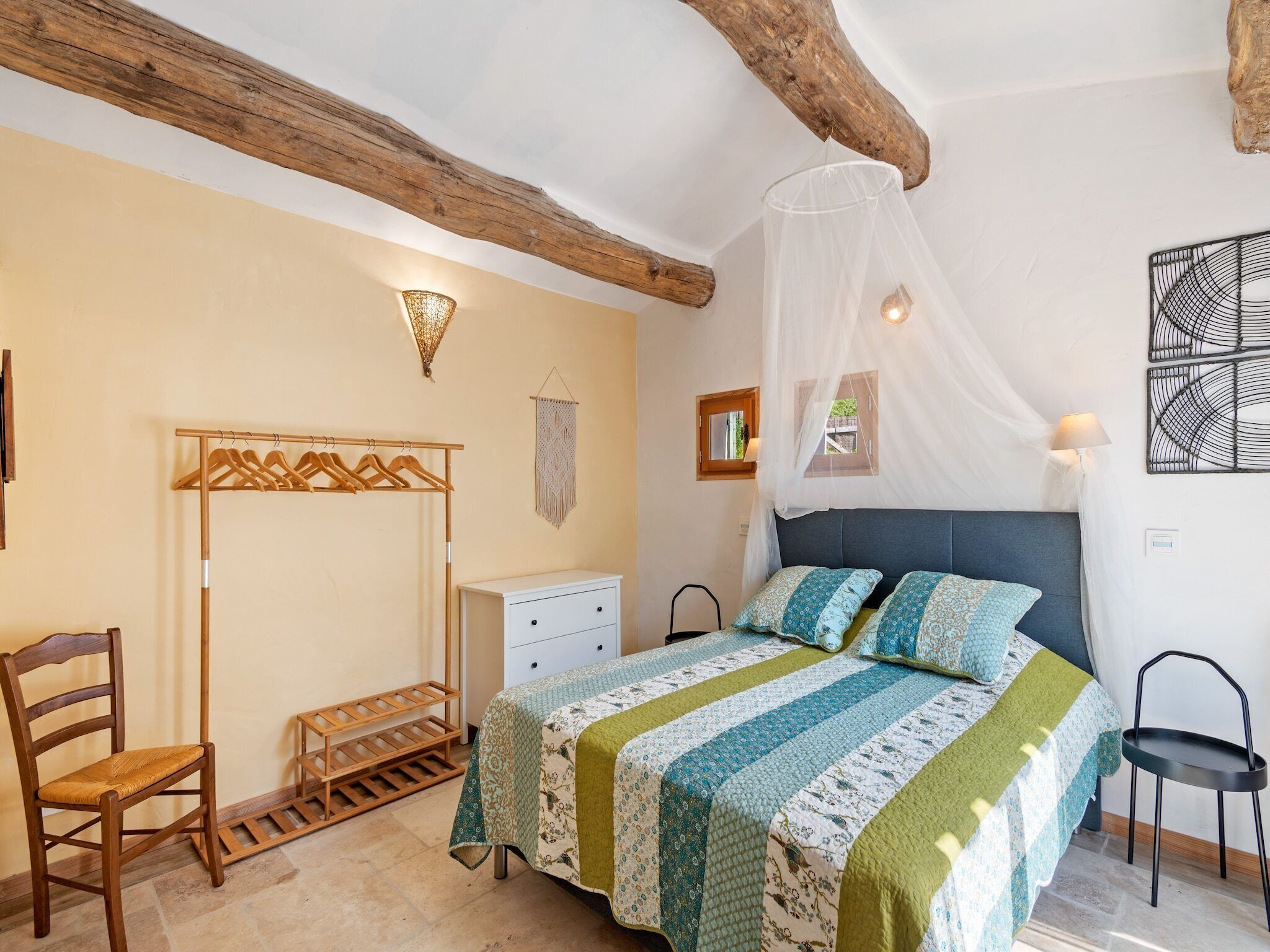A bedroom with a bed, a wooden chair, a small table, a wardrobe, and a window with a view.