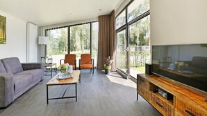 Un salon moderne avec un canapé gris, une table basse en bois, deux fauteuils, un téléviseur posé sur un meuble en bois et de grandes fenêtres avec des rideaux.