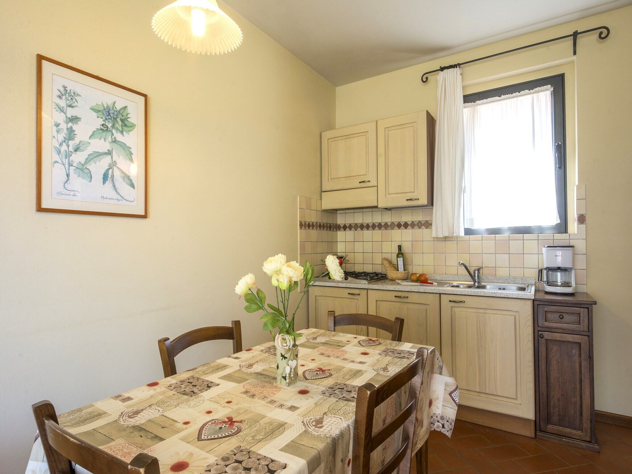 A kitchen with a dining table, chairs, a hanging light fixture, a framed botanical illustration, and a window with curtains.