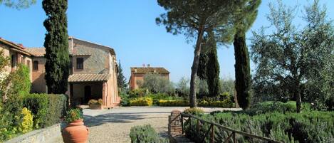 Una villa rústica con fachada de piedra, un camino de grava que conduce a un puente de madera, y un jardín bien cuidado con cipreses y lavanda.