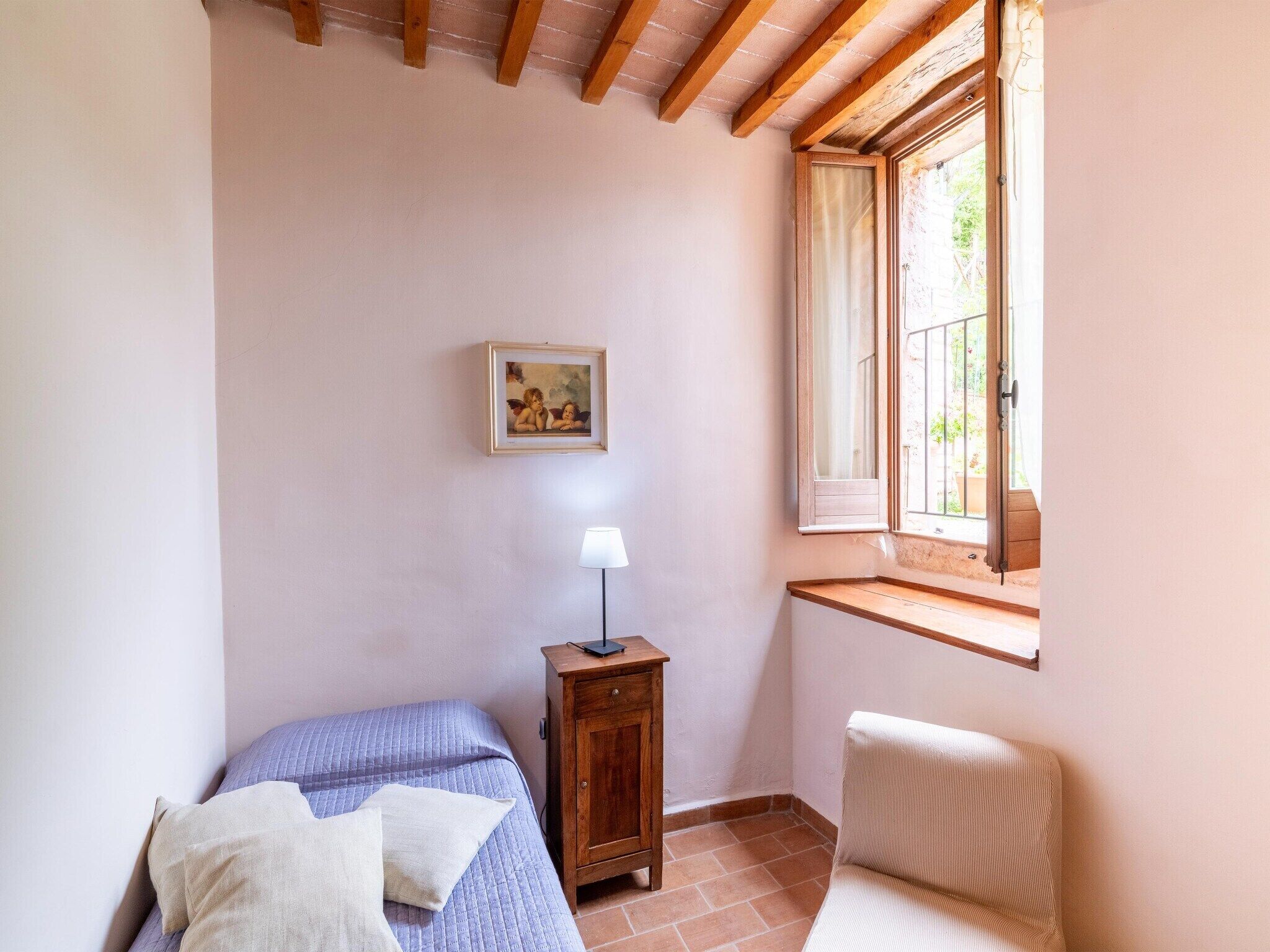 A bedroom with a bed, a nightstand, a lamp, a chair, a window, and a painting on the wall.