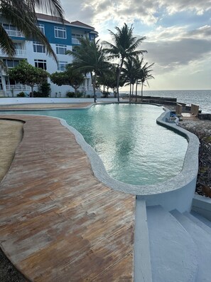 A swimming pool with a wooden deck, surrounded by palm trees and a building with balconies. - Luxury penthouse Ocean view (Tower Isle)