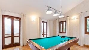 Une table de billard dans une pièce dotée d’un plancher en bois, de grandes fenêtres et d’un luminaire au plafond.