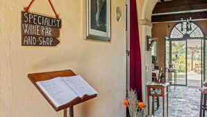 Un letrero de madera con la inscripción 'Specialist Wine-Bar and Shop', un atril con menú, y la entrada a un edificio con una cortina roja y vistas al exterior.