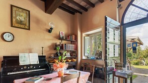 Una habitación acogedora con un piano, una mesa cubierta con un mantel floral, una silla, una estantería y una ventana con vistas a la vegetación.