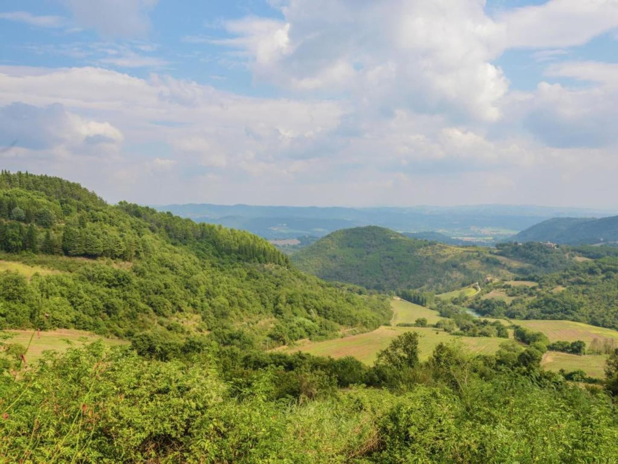 Vistesa panoràmica d'una vall verda i exuberant amb turons ondulants i muntanyes llunyanes sota un cel amb núvols dispersos.