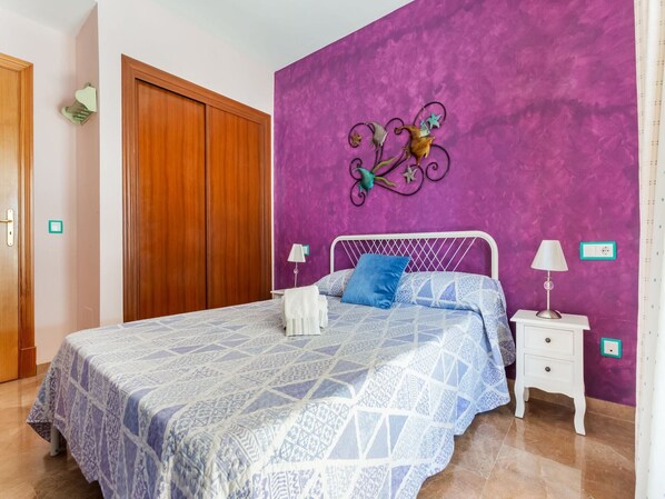 Un dormitorio con cama, mesitas de noche, una pared morada y una puerta de madera.