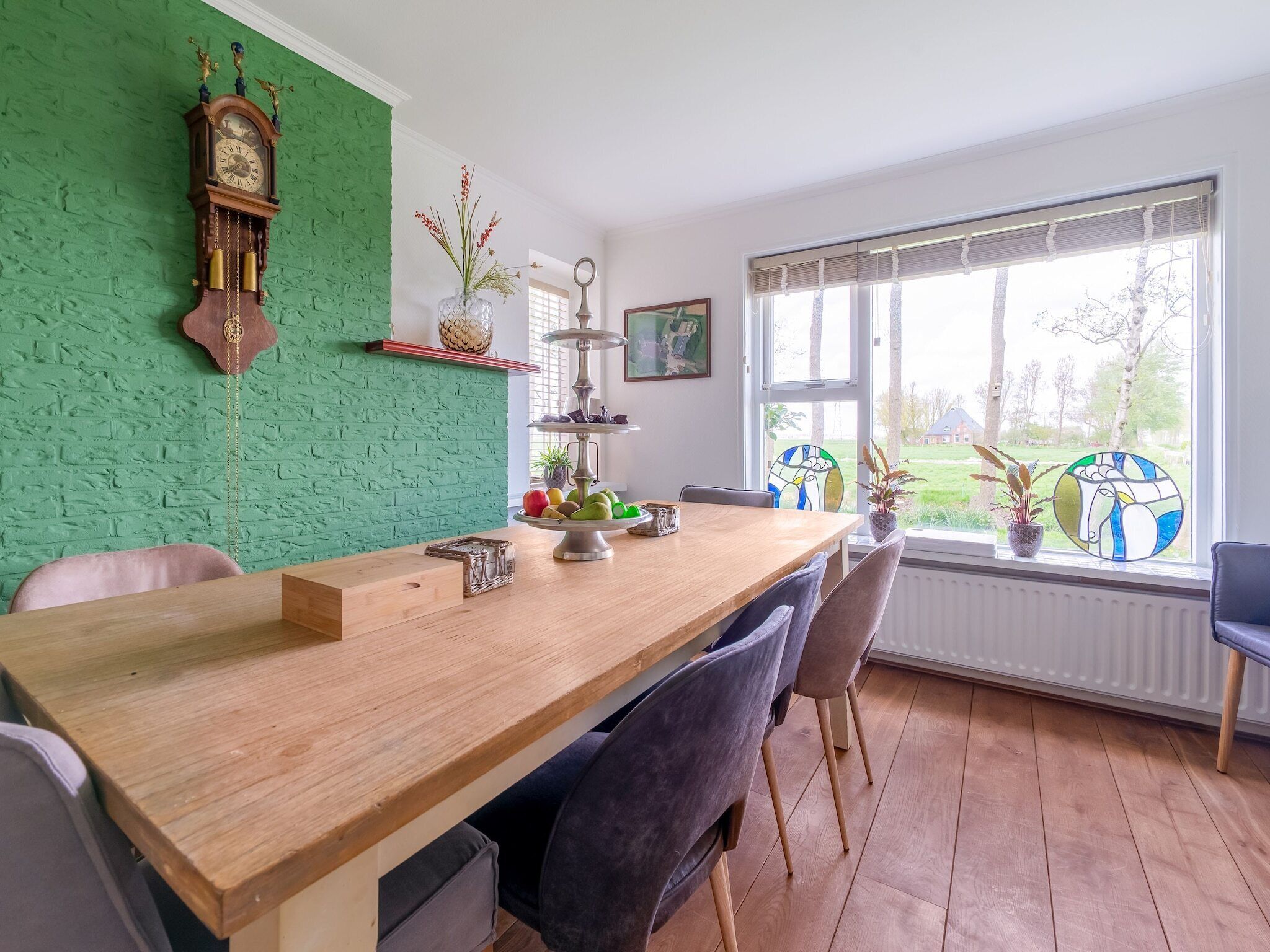 Ein Essbereich mit einem Holztisch, Stühlen, einer Wand mit Grüner Struktur, einer Uhr, einer Vase mit Blumen und einem Fenster mit Blick auf Bäume.