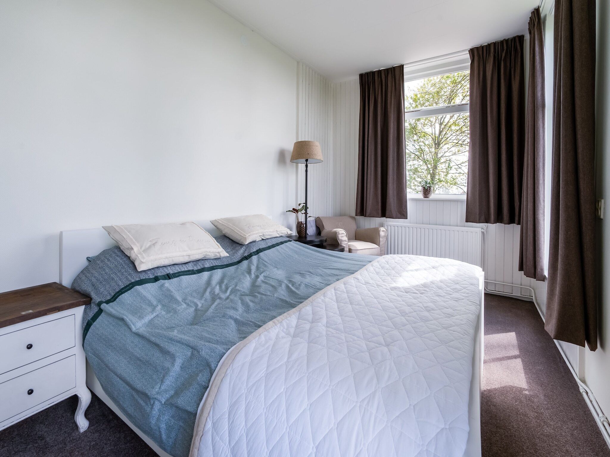 A bedroom with a bed, nightstand, chair, lamp, and window with curtains.