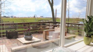 Un espace patio offrant une vue sur un paysage rural, comprenant un champ et un bâtiment situé au loin. On y trouve des plantes en pot et un banc en bois.