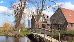 Une maison traditionnelle néerlandaise avec un pont en bois enjambant un canal, entourée d’arbres et d’un champ verdoyant.