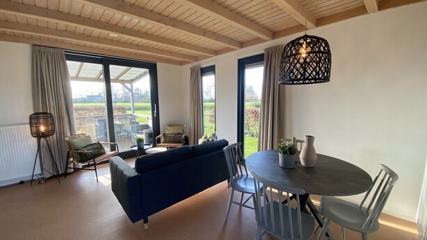 Uma sala de estar moderna com um sofá azul, mesa de jantar e cadeiras. Há uma janela grande com cortinas, um abajur e uma luminária pendente.