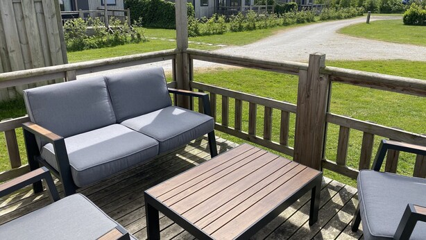 A wooden deck with a grey sofa and a table, overlooking a gravel path and a row of houses. - Chalet in Nature, Friesland (De Veenhoop)
