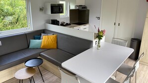 Een compact keukenhoekje met een magnetron, fornuis en gootsteen. Er is een eettafel met stoelen, een klein rond tafeltje en een bank met kussens. Een raam zorgt voor natuurlijk licht, en op de tafel staat een vaas met bloemen.