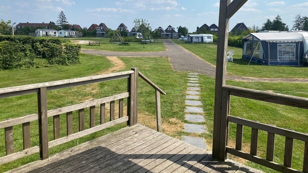 Een houten terras loopt over in een gazon met een duidelijke pad, een overdekte picknickplaats en een caravan in de verte.