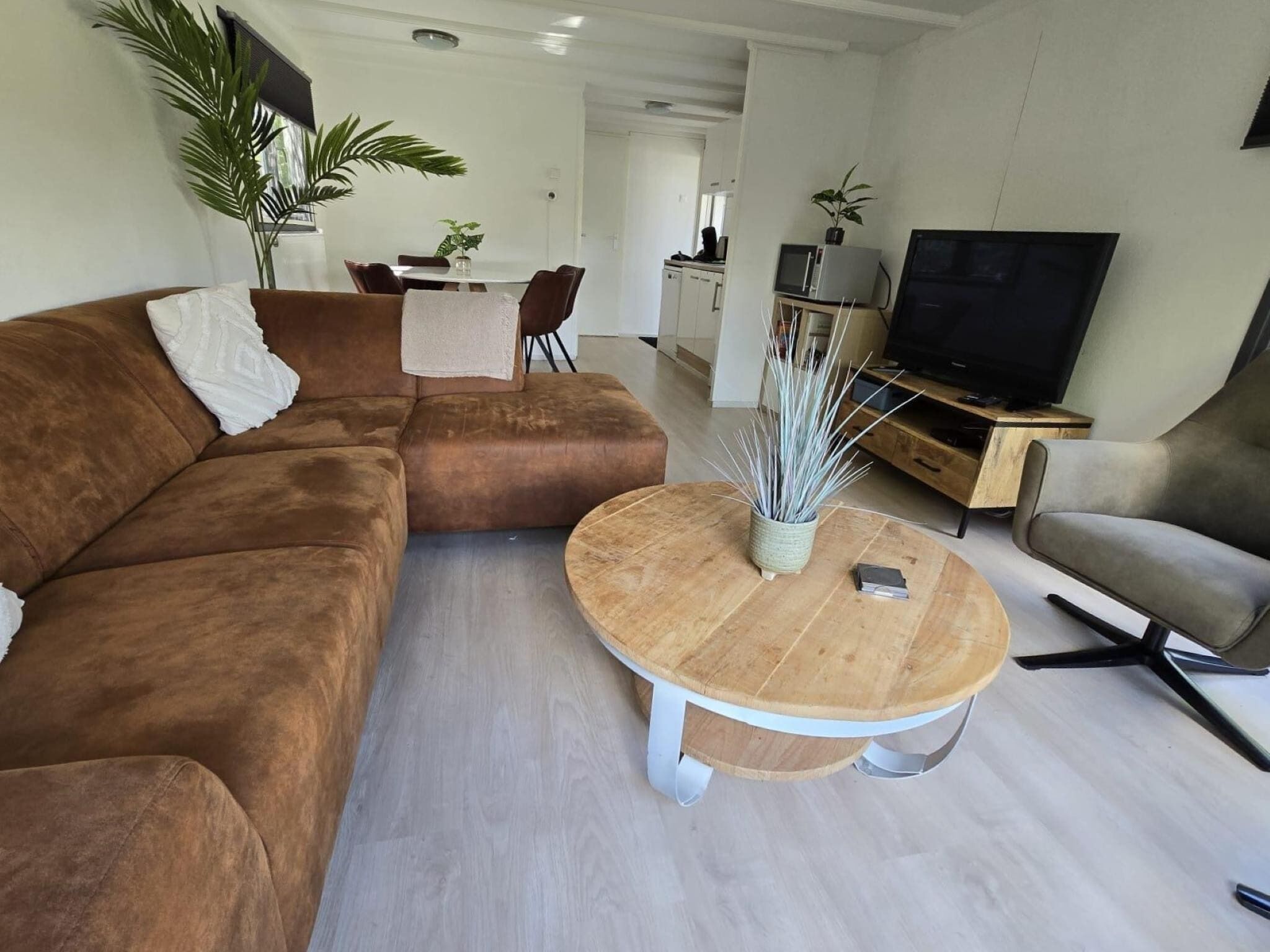 A modern living room with a brown sectional sofa, a round wooden coffee table, a TV, and a kitchen in the background.