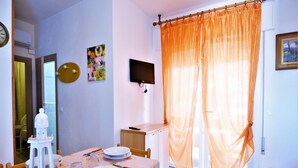 A dining area with a table set for four, a television mounted on the wall, and a window with orange curtains. - Apartment in Rosolina Mare near the Sea (Rosolina Mare (RO))
