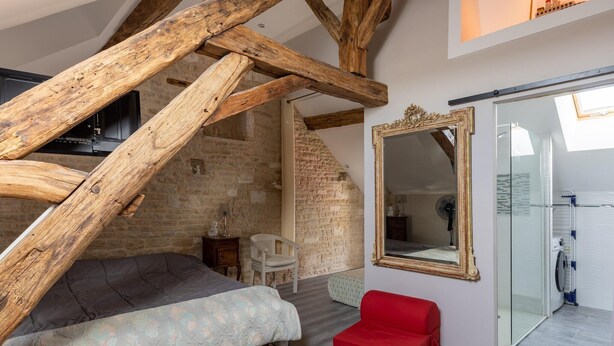 A room with exposed wooden beams, a bed, a red chair, a large mirror, and a glass door leading to a bathroom.