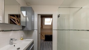 A bathroom with a white sink, mirror, and shower. There is a skylight above the shower and a wooden door leading to another room.
