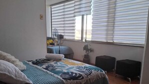 A bedroom with a bed, a bedside table, a chair, a window with blinds, and a wooden floor.