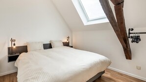 Un dormitorio con techo inclinado, tragaluz, cama con ropa de cama blanca, dos mesitas de noche con lámparas y estructura de vigas de madera.