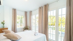 Un dormitorio con una cama grande, una silla, un escritorio con un florero y ventanas amplias con cortinas.